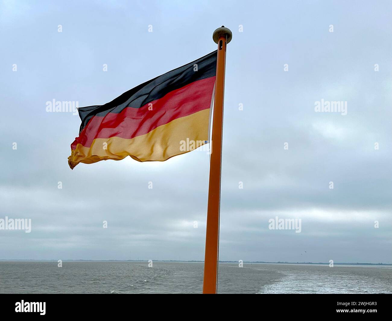 Eine deutsche Flagge weht an einem kalten Wintertag in der Nordsee zwischen dem turbulenten Meer und dem bewölkten Himmel an Bord der Fähre nach Langeoog Island Stockfoto