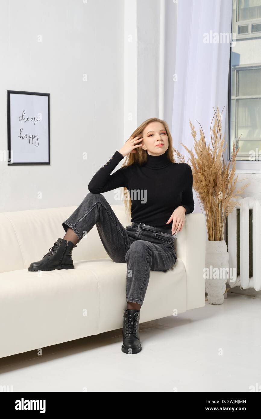 Niedliche Frau in schwarzem Freizeitoutfit sitzt auf einem weißen Ledersofa Stockfoto