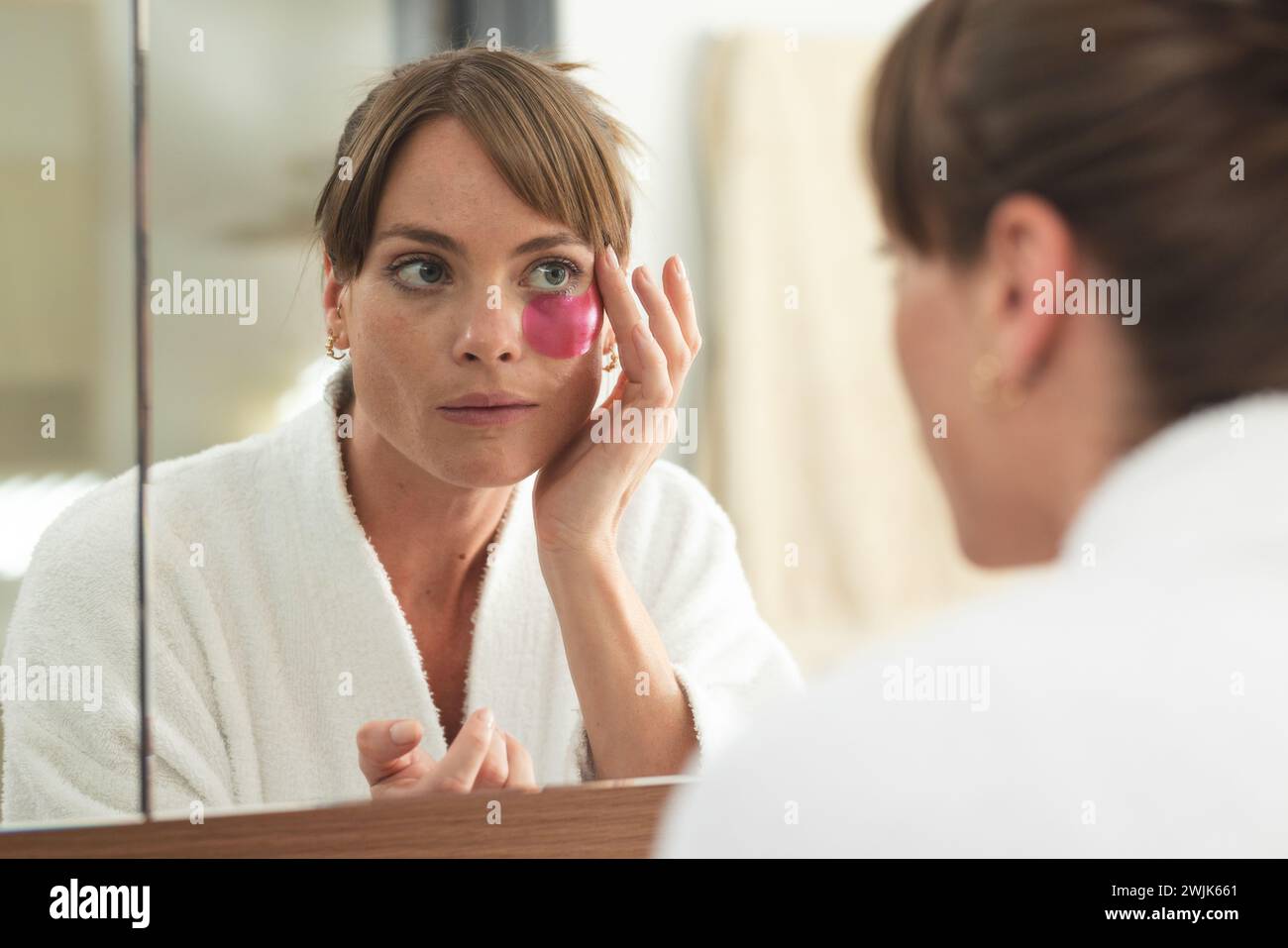 Eine kaukasische Frau untersucht ihr Gesicht im Spiegel und trägt eine rosa Augenmaske auf Stockfoto