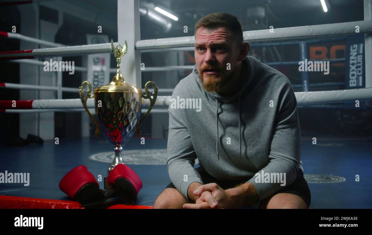 Männlicher Boxer sitzt in der Nähe des Boxrings und spricht mit dem Interviewer über den Sieg im Turnier. Athlet spricht nach Meisterschaft oder Wettkampf im Boxstudio. Eine Tasse Sieger und Boxhandschuhe liegen in der Nähe des Käufers. Stockfoto