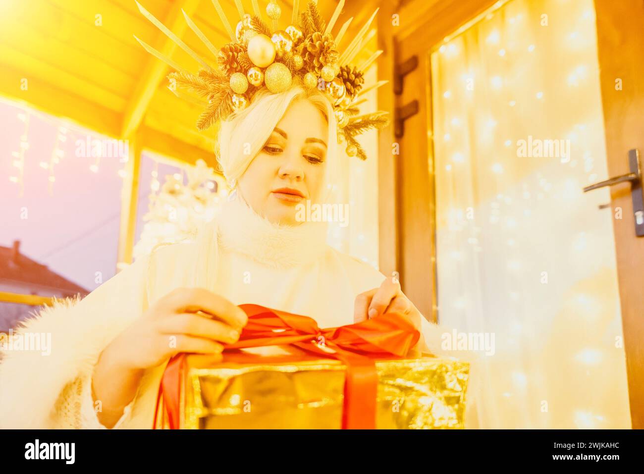 Eine Frau in einem weißen Kleid hält eine goldene Box mit einem roten Band. Sie trägt eine Krone auf dem Kopf. Die Frau scheint die Geschenkbox zu öffnen. Stockfoto