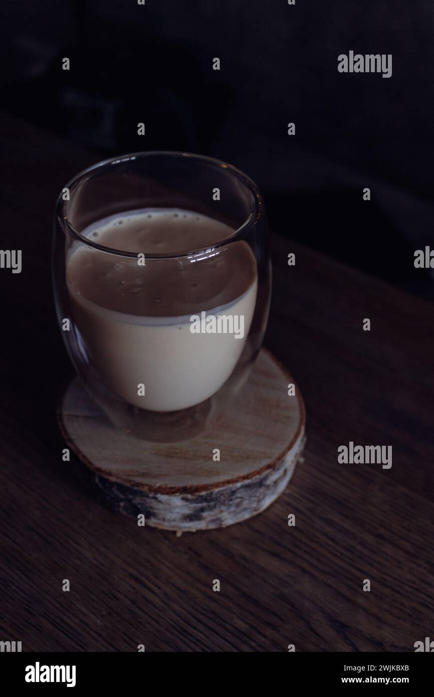 Cremiger Kaffee auf Holzständer auf dunklem Hintergrund. Milchschaum mit Espresso im Glas. Frappe in Glas auf dem Tisch. Kalter Cappuccino im Becher. Schlagsahne. Stockfoto