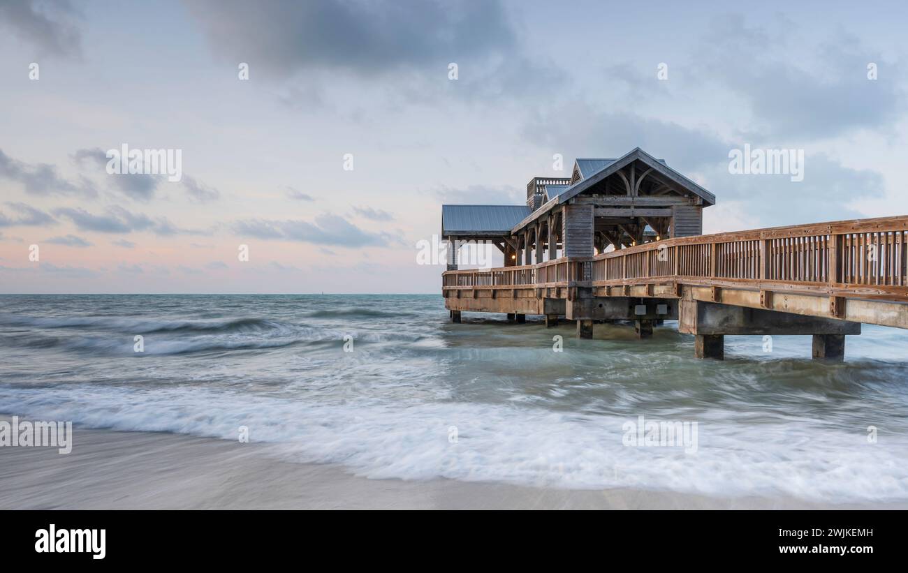 Holzpier in Key West, Florida, der bei Sonnenaufgang in das ruhige tropische Wasser des Ozeans reicht. Es gibt einen Hauch von Orange am Himmel Stockfoto