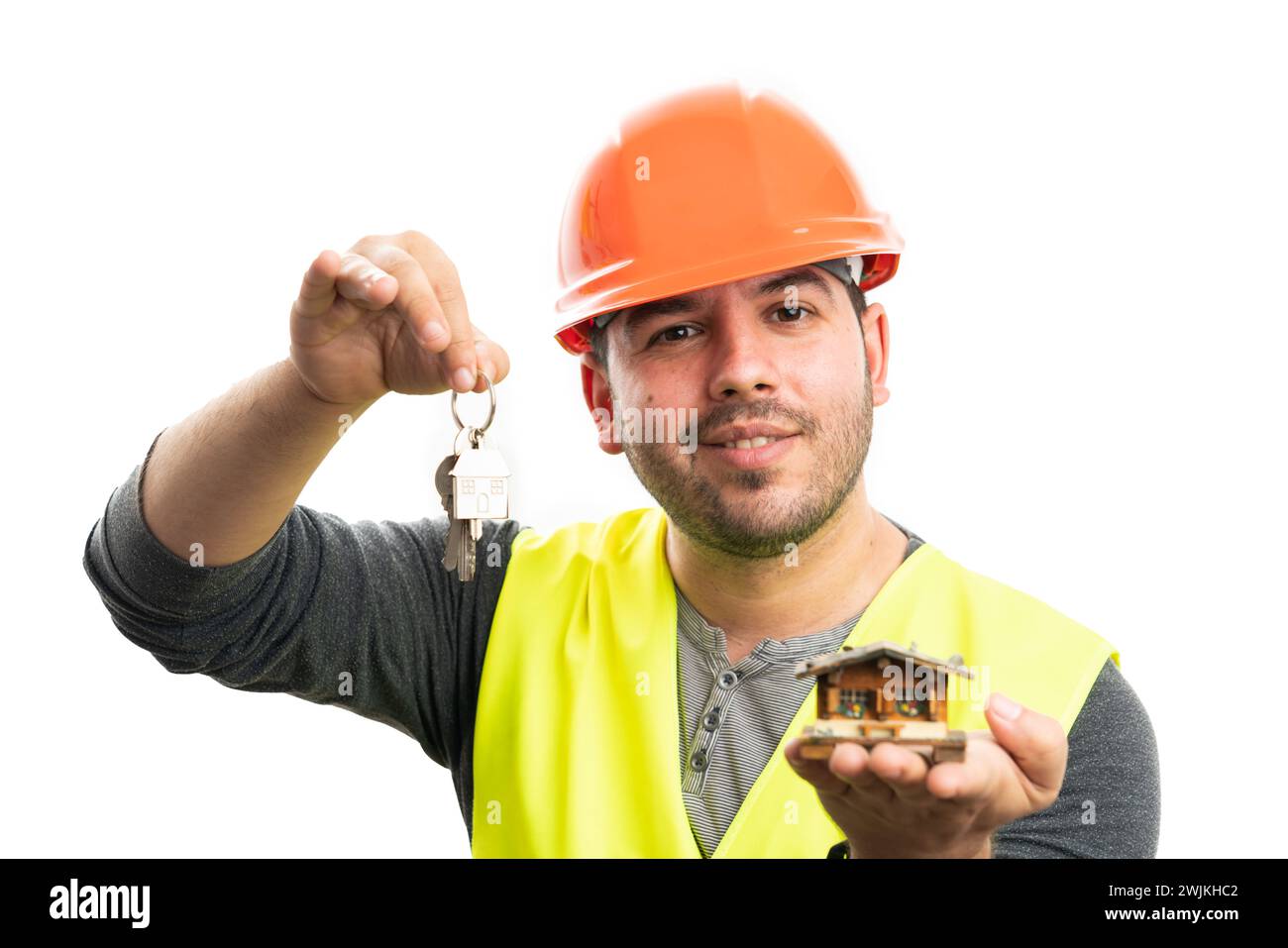 Lächelnder erwachsener Baumeister-Mann mit orangefarbenem Schutzhelm und gelber Weste, der Miniatur-Holzhaus mit Hauptschlüsseln auf weißem Hintergrund präsentiert Stockfoto