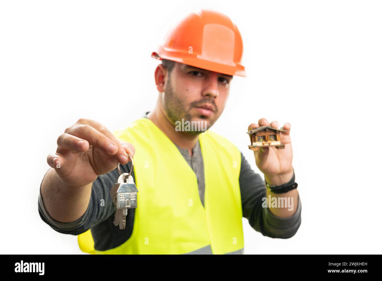 Männlicher Baumeister mit orangefarbenem Schutzhelm, der ein hölzernes Miniaturhaus hält, bietet Schlüssel aus der Nähe selektiven Fokus auf weißem Hintergrund Stockfoto