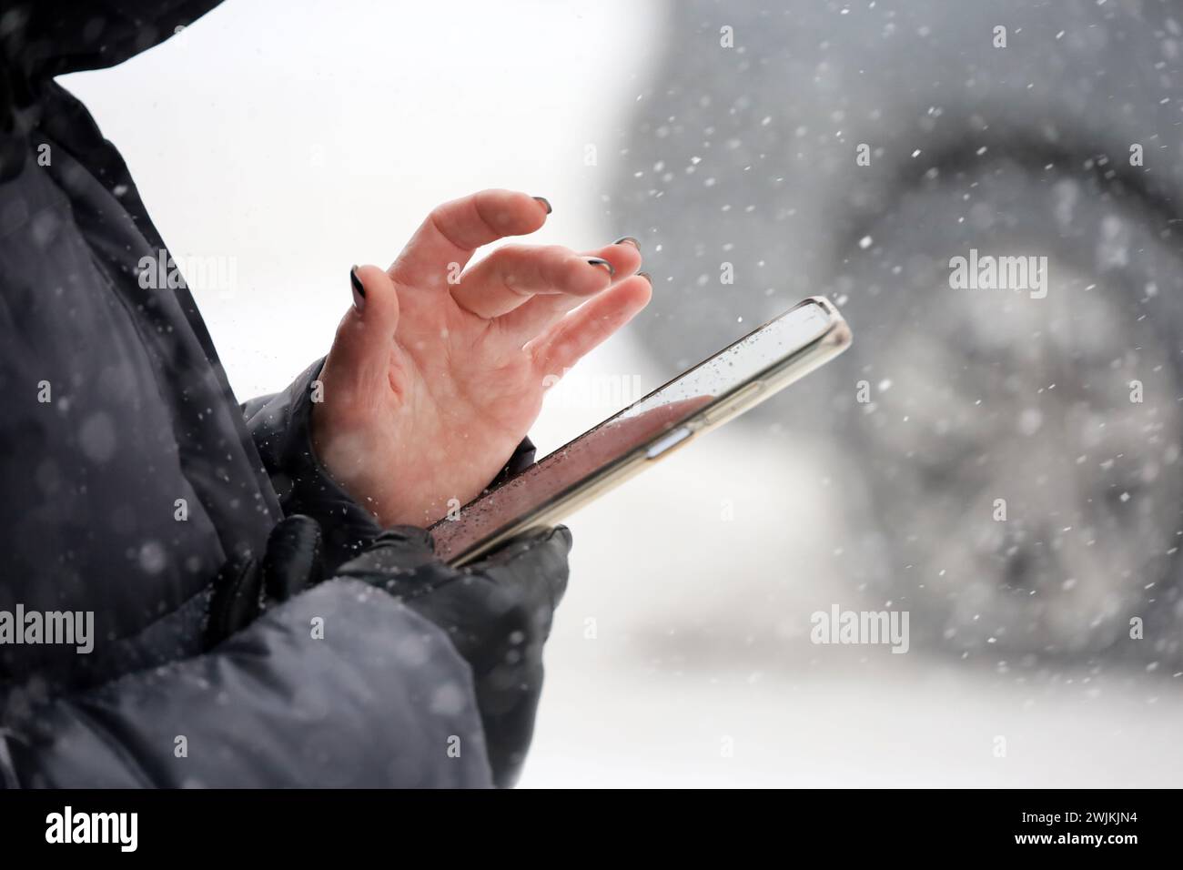 Weibliche Hände mit Smartphone nähern sich verschwommenem Fahrzeughintergrund bei Schneefall. Frau, die im Winter auf einer Straße in der Stadt ein Handy benutzt Stockfoto