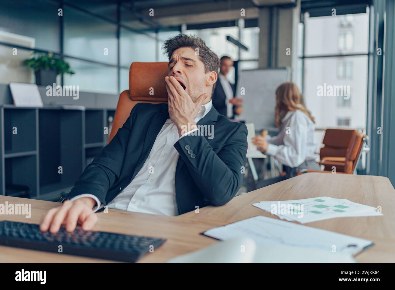 Junger übermüdeter Geschäftsmann gähnt, während er in einem modernen Büro mit Computer arbeitet Stockfoto
