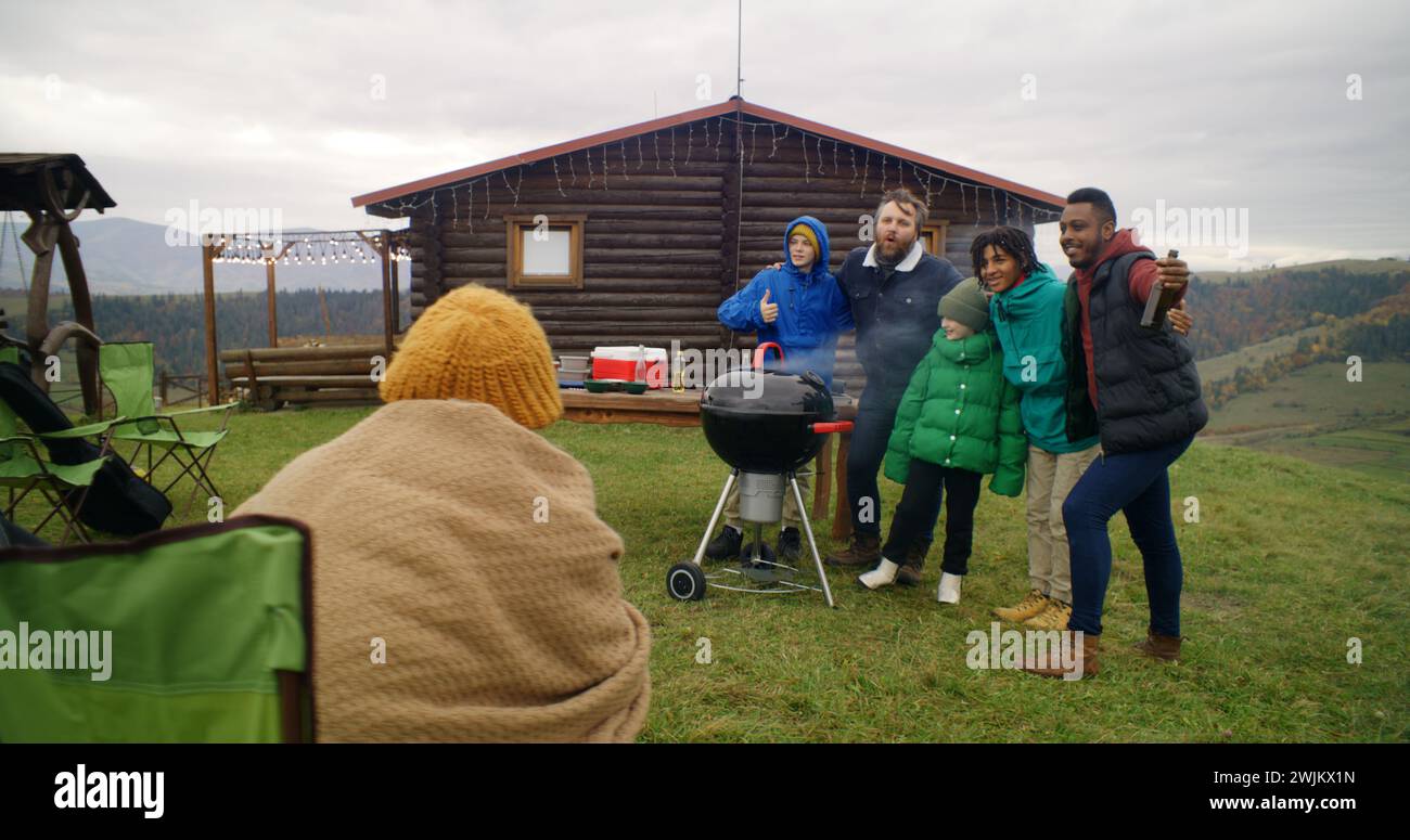 Eine multiethnische Gruppe von Reisenden steht neben dem Grillgrill und posiert vor der Kamera. Frau im Lagerstuhl macht Fotos am Telefon. Große Familie, die vor dem Ferienhaus in den Bergen ruht. Tourismus und Reisen. Stockfoto