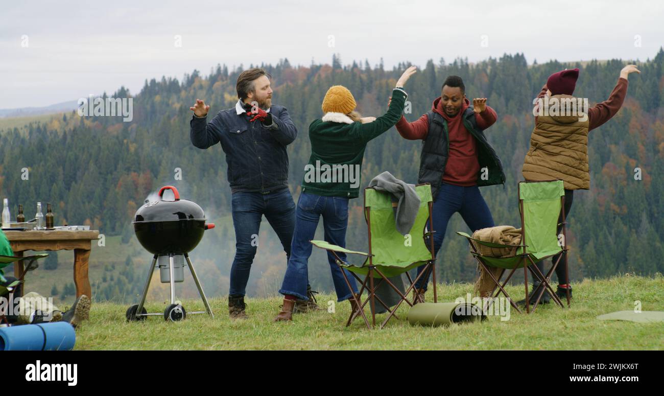 Gruppe von Reisenden hat Spaß beim Barbecue auf einem Hügel. Fröhliche multiethnische Freunde tanzen während der Urlaubsreise im Freien. Berglandschaft und Wald im Hintergrund. Outdoor-Enthusiasten. Stockfoto