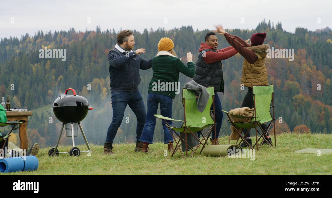 Gruppe von Reisenden hat Spaß beim Barbecue auf einem Hügel. Fröhliche multiethnische Freunde tanzen während der Urlaubsreise im Freien. Berglandschaft und Wald im Hintergrund. Outdoor-Enthusiasten. Stockfoto