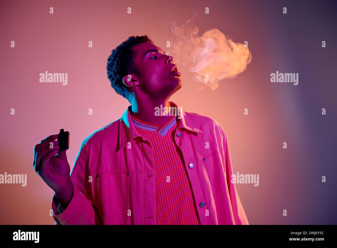 Porträt eines afroamerikanischen Mannes, der Rauch ausatmet, während er eine Zigarette auf buntem Hintergrund hält Stockfoto