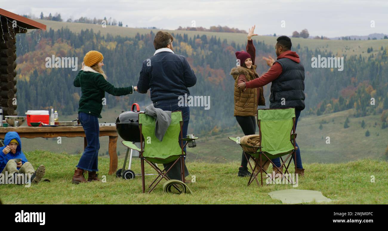 Gruppe von Reisenden hat Spaß beim Barbecue auf einem Hügel. Fröhliche multiethnische Freunde tanzen während der Urlaubsreise im Freien. Berglandschaft und Wald im Hintergrund. Outdoor-Enthusiasten. Stockfoto
