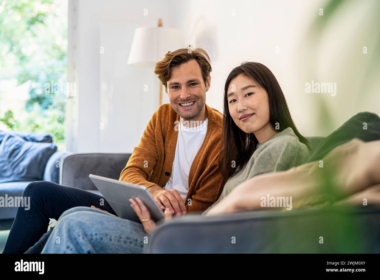 Erwachsenes Paar, das mit einem digitalen Tablet auf dem Sofa sitzt und auf die Kamera blickt Stockfoto