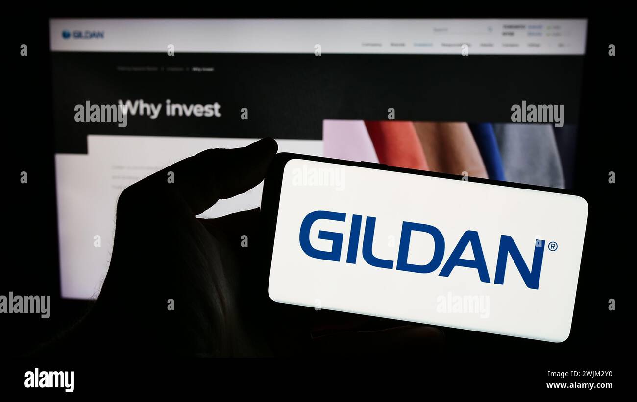 Person, die ein Mobiltelefon mit dem Logo des kanadischen Bekleidungsunternehmens Gildan activewear Inc. Vor der Business-Website hält. Schwerpunkt auf dem Display des Telefons. Stockfoto