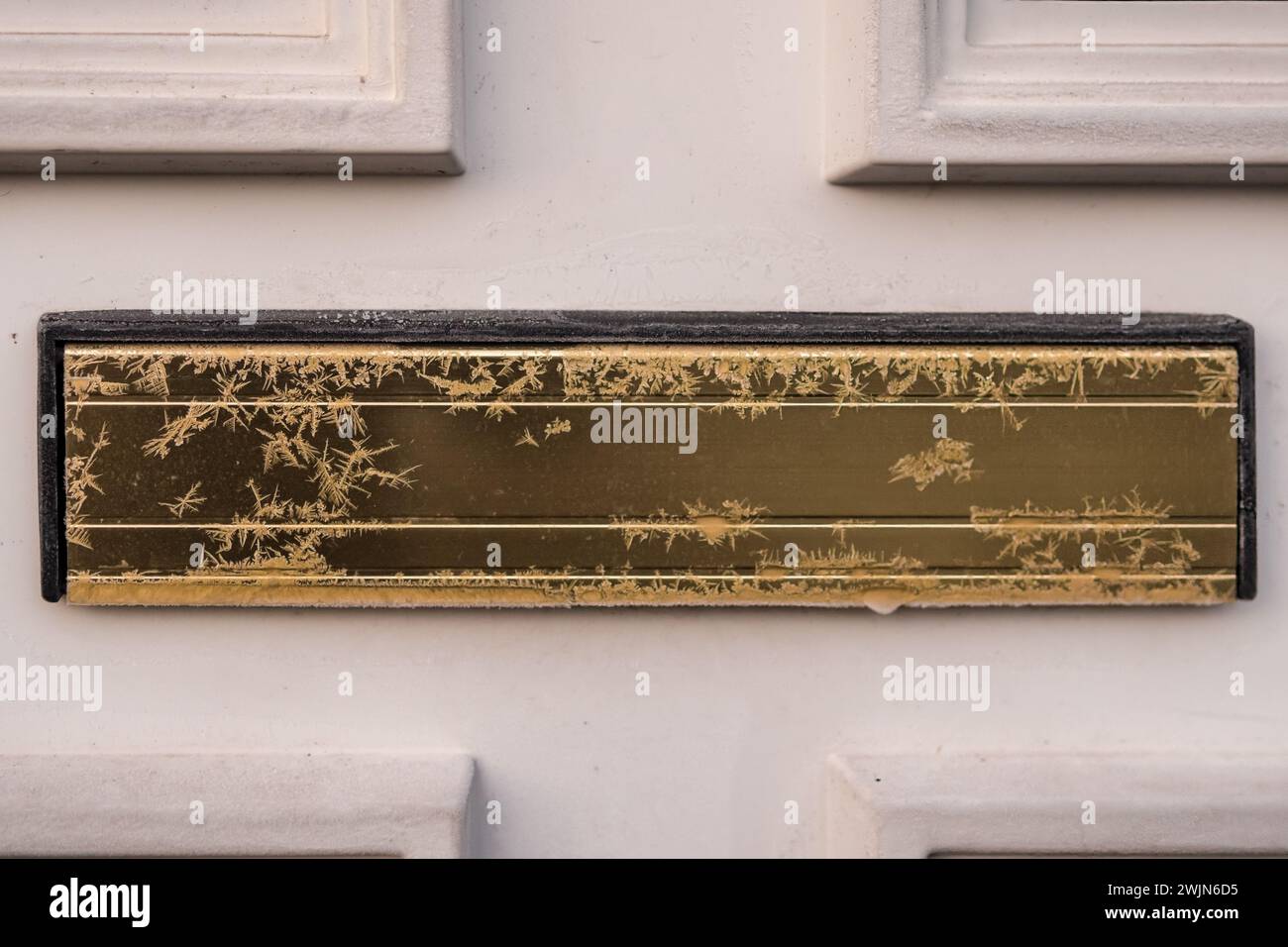 Frost-/Eismuster auf einem goldfarbenen Briefkastendeckel in der weißen Tür. Stockfoto