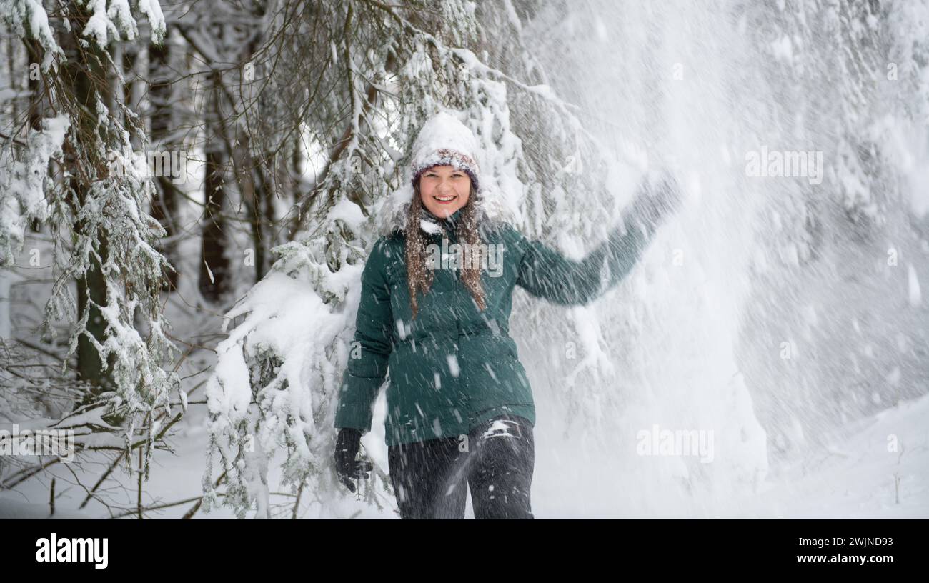Eine fröhliche Frau in einem grünen Wintermantel, ihr Lachen ertappt in einem Schneespiel von einem Baum. Sie trägt einen schneebedeckten Hut und den Wald Stockfoto