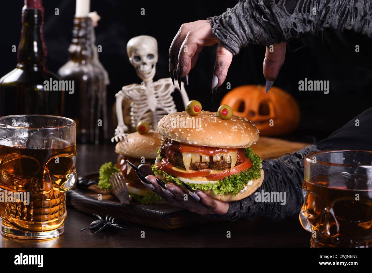 Die Hexenhände halten einen Monster Burger in ihren Händen auf einem sitzenden Skelett. Die perfekte Vorspeise für die Halloween Party Stockfoto