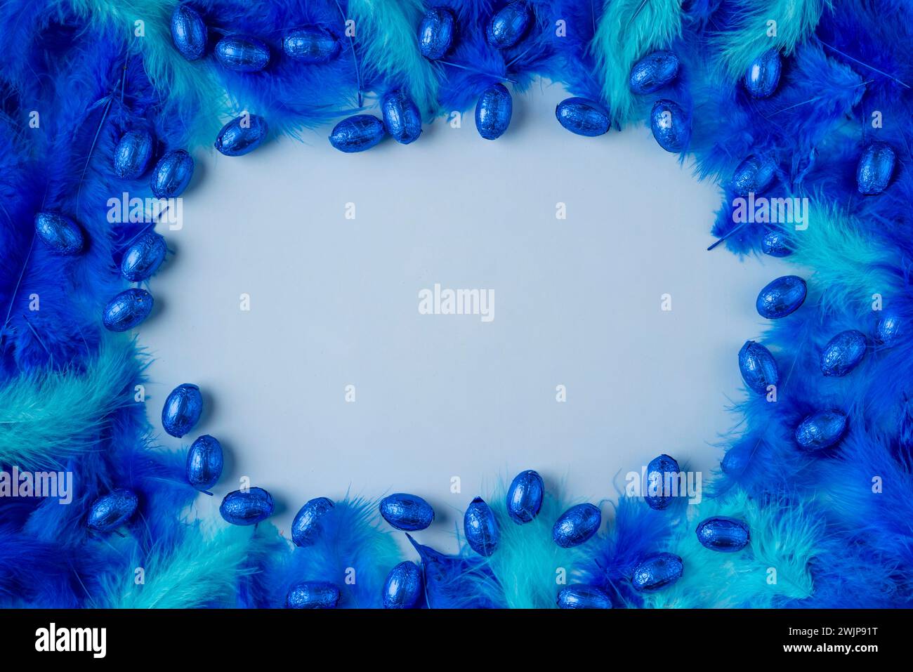 Rahmen aus blauen und türkisfarbenen Federn mit blauen Schokoladeneiern auf hellblauem Hintergrund Stockfoto