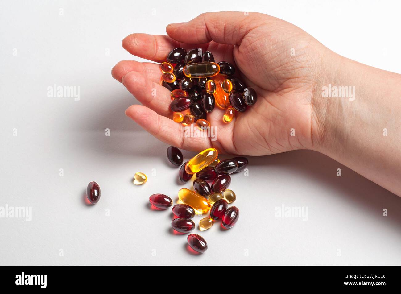 Eine Person Hand zeigt eine Sammlung von verschiedenen farbigen und geformten Nahrungsergänzungskapseln, wobei einige auf eine weiße Oberfläche verschüttet werden. Stockfoto