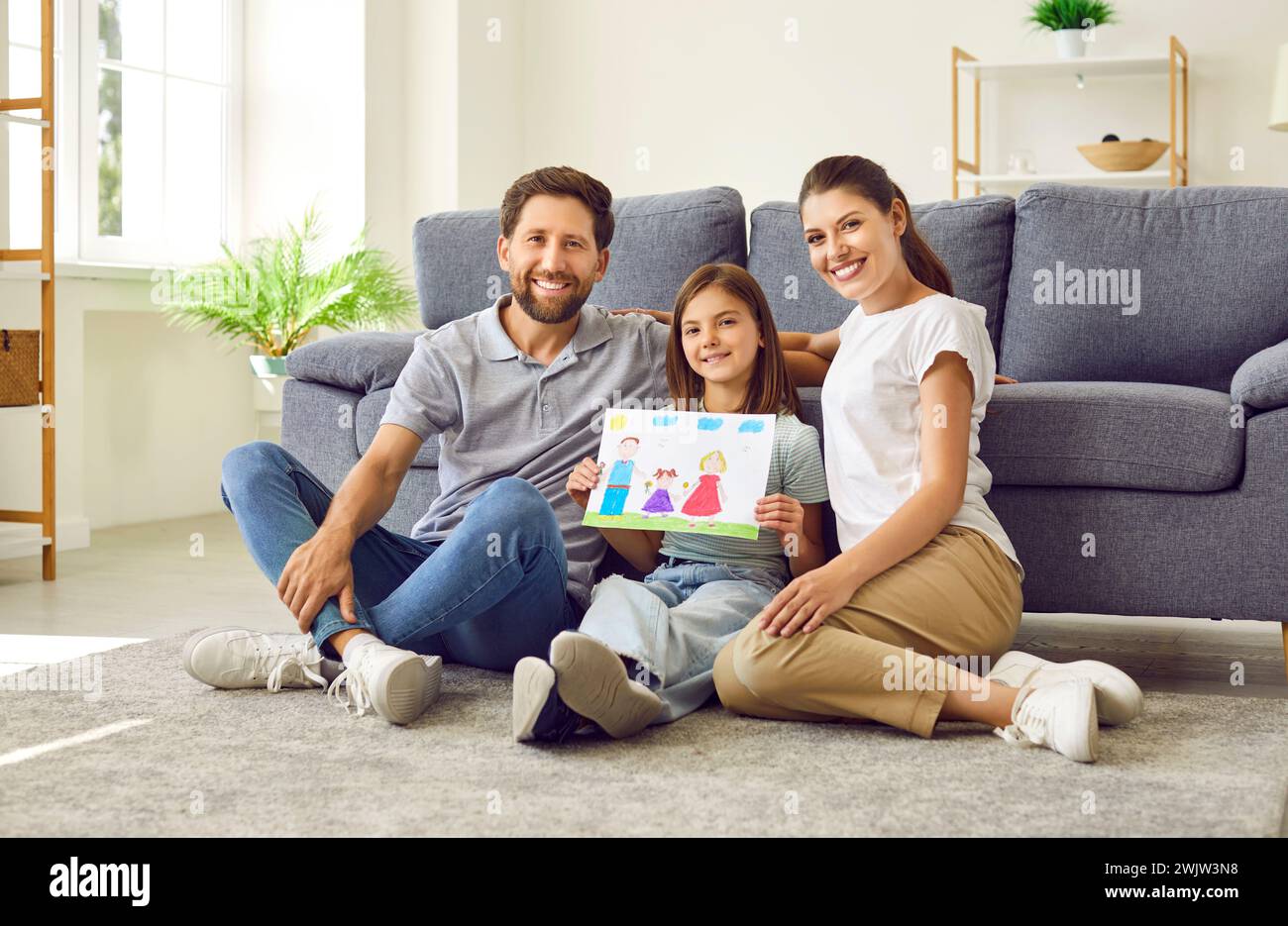 Fröhliches kleines Mädchen, das mit ihren jungen Eltern zu Hause sitzt und eine Zeichnung einer glücklichen Familie hält. Stockfoto