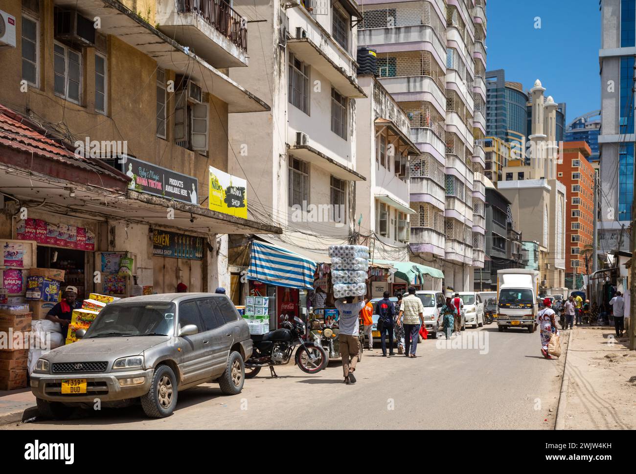 Ein Portier trägt Toilettenrollen auf dem Kopf, an Geschäften vorbei, in dieser allgemeinen Sicht des Lebens in einer Straße im Zentrum von dar es Salaam, Tansania. Stockfoto
