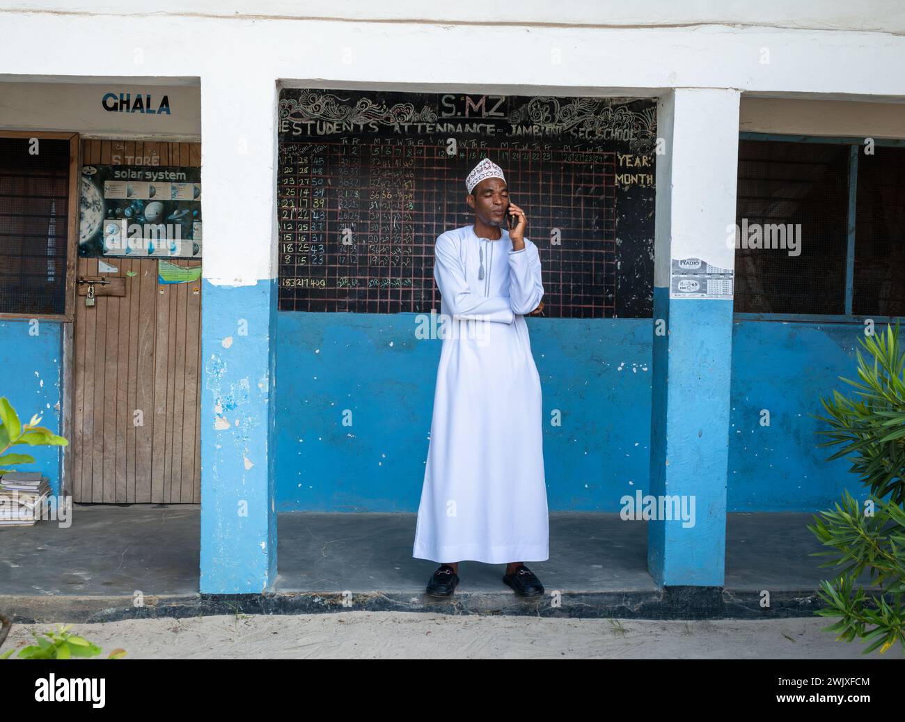 Ein männlicher Lehrer trägt ein langes Kanzu-Gewand und eine koffiabestickte Kappe und benutzt ein Handy an der Jambiani Secondary School in Sansibar, Tansania. Stockfoto