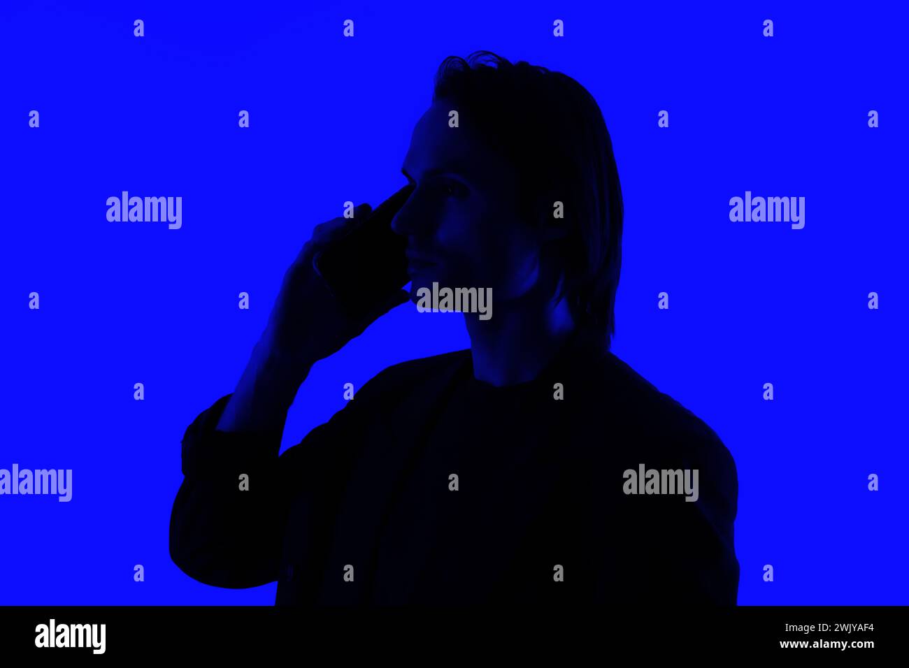 Unerkennbare Silhouette ein eleganter Geschäftsmann, der auf einem Smartphone spricht, in einer Jacke, die auf einem blau leuchtenden Hintergrund steht, ein futuristisches Konzept Stockfoto