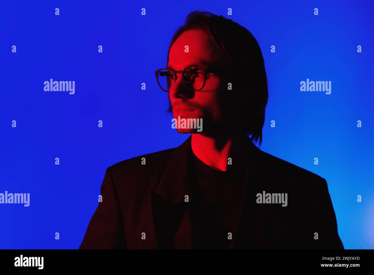 Eleganter Geschäftsmann in Brille in einer Jacke, der in einem dunklen Raum mit leuchtendem Red Neon Gllowing Light auf dem Gesicht auf blauem Hintergrund steht Stockfoto