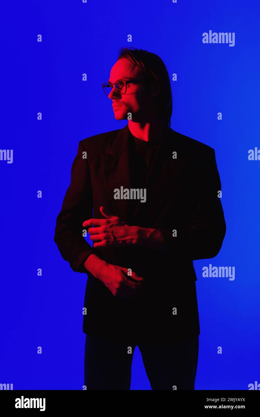 Eleganter Geschäftsmann in Brille in einer Jacke, der in einem dunklen Raum mit leuchtendem Red Neon Gllowing Light auf dem Gesicht auf blauem Hintergrund steht Stockfoto