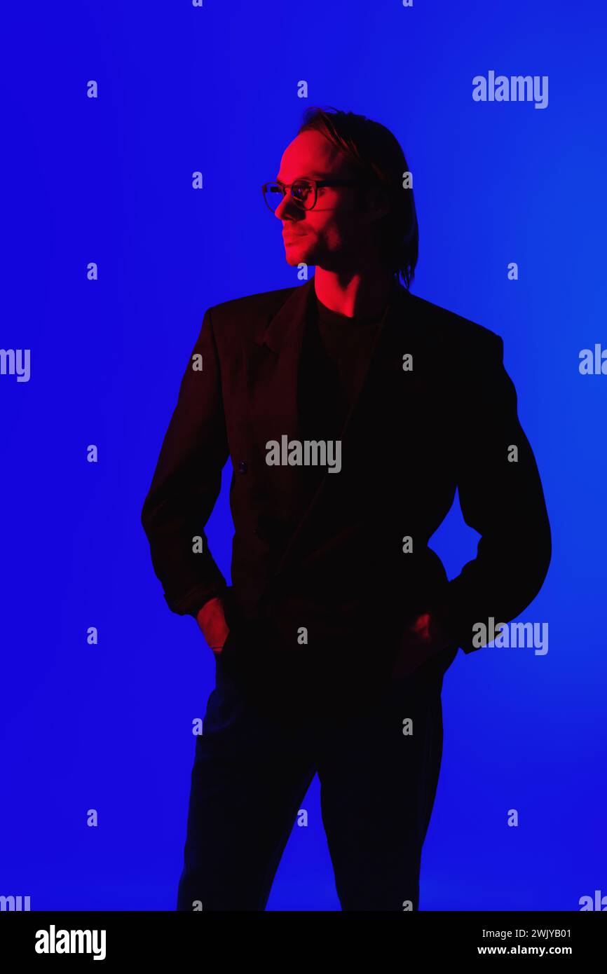 Eleganter Geschäftsmann in Brille in einer Jacke, der in einem dunklen Raum mit leuchtendem Red Neon Gllowing Light auf dem Gesicht auf blauem Hintergrund steht Stockfoto