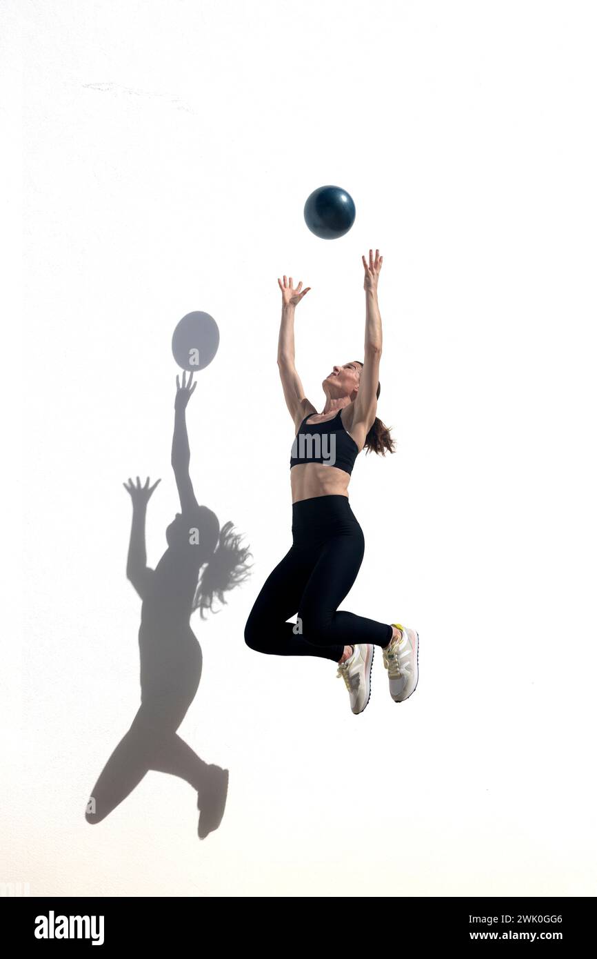 Frau, die in die Luft springt, um einen Ball zu fangen, draußen vor einer weißen Wand mit Schatten. Stockfoto