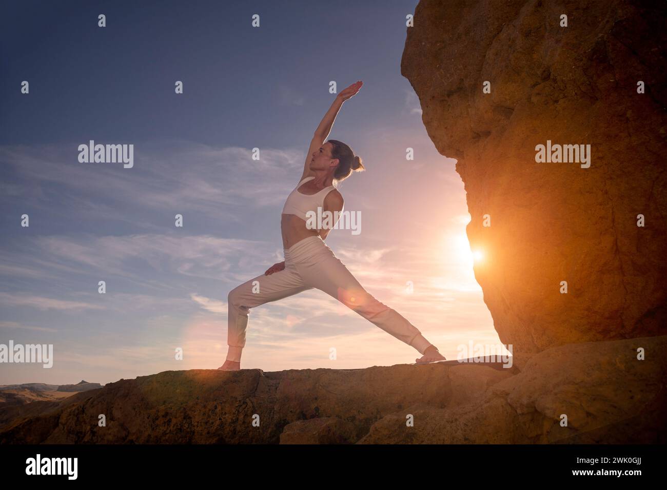 Frau, die bei Sonnenaufgang auf Felsen steht, Sonnenuntergang, die eine umgekehrte Krieger-Yoga-Pose macht. Stockfoto
