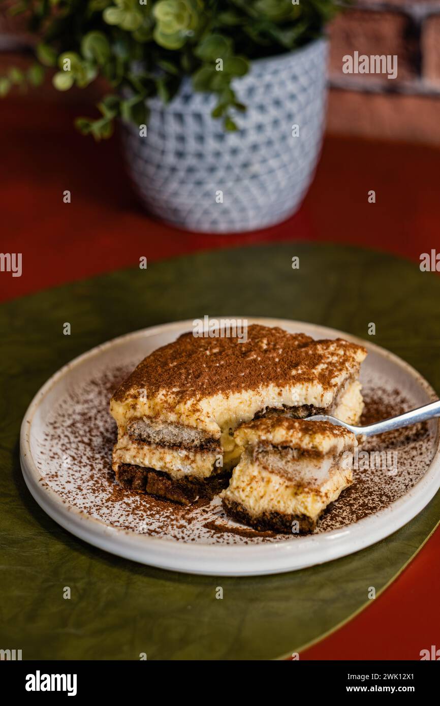 Nahaufnahme einer Portion Tiramisu-Kuchen mit Kakaopulver, serviert mit einer Gabel auf einem weißen Teller. Grüne Pflanze in einer Vase auf dem Restauranttisch, b Stockfoto