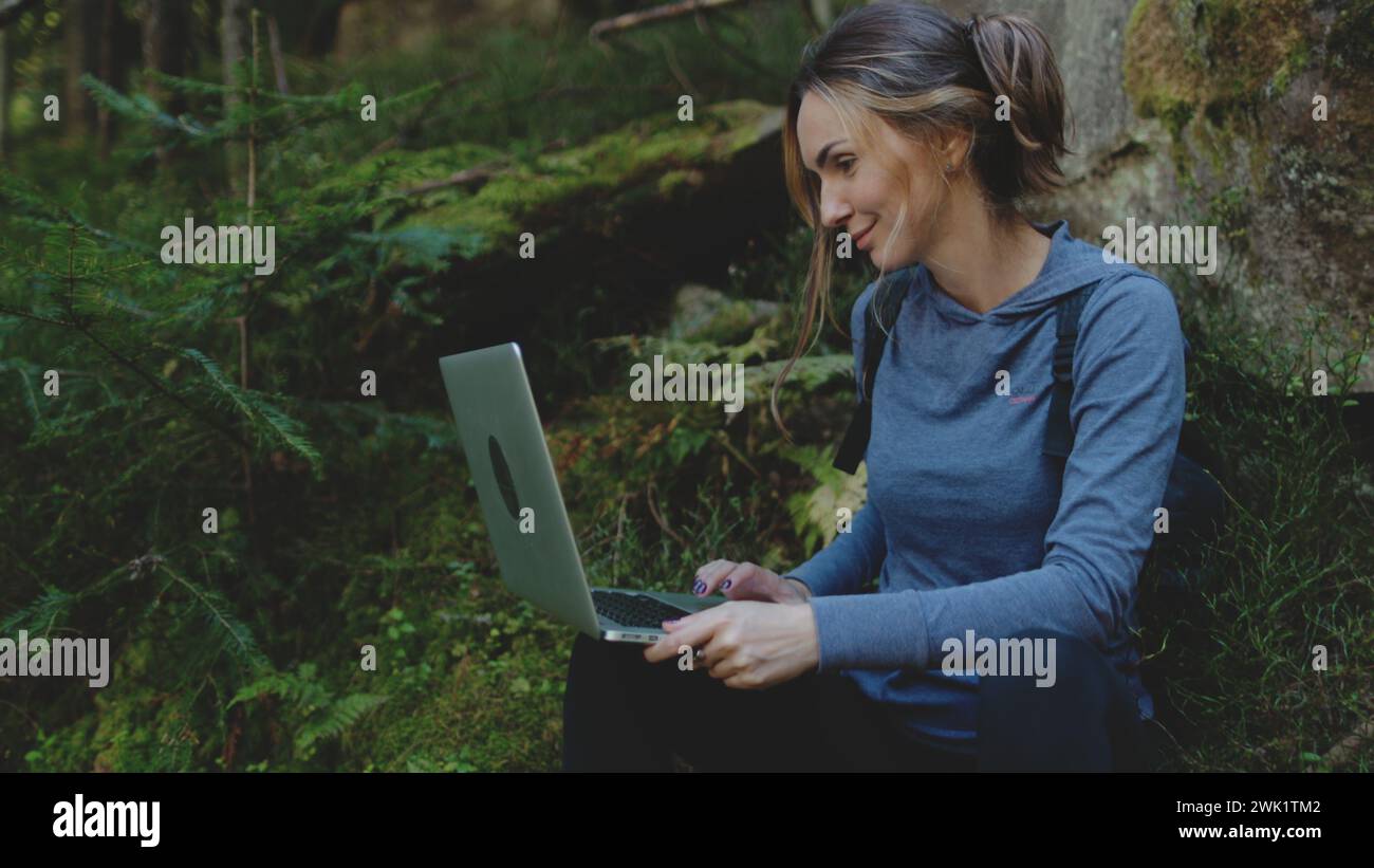 Frau, die auf einem Laptop sitzt, sitzt unter dem Stein im grünen Wald. Konzept der Technologie Online-digitalen Nomaden-Berufsausbildung für moderne Menschen. Weißes Mädchen, das online arbeitet Stockfoto