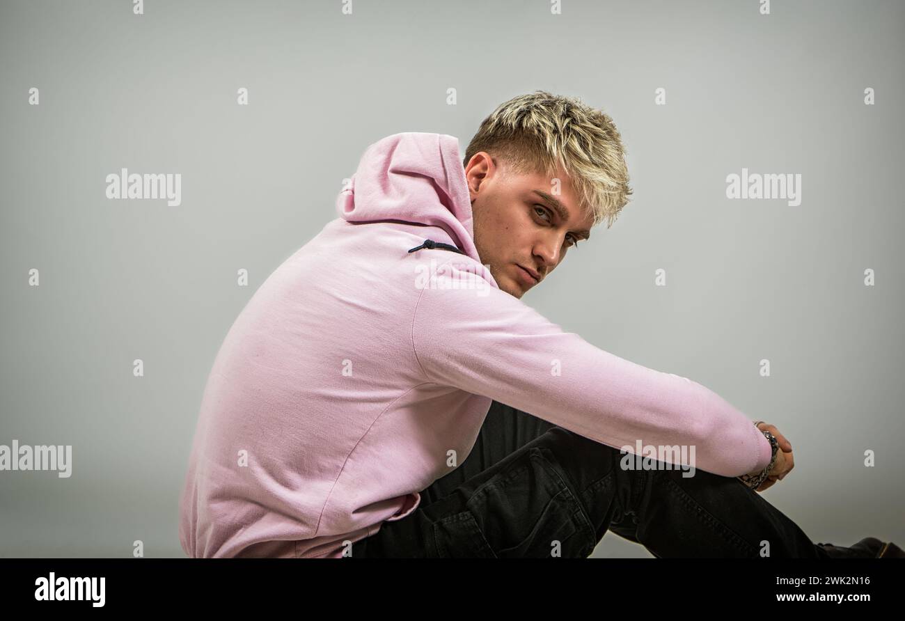 Ein Mann mit einem rosa Hoodie sitzt auf diesem Foto auf dem Boden. Er wirkt entspannt und gedankenlos, mit gekreuzten Beinen und aufliegenden Händen Stockfoto