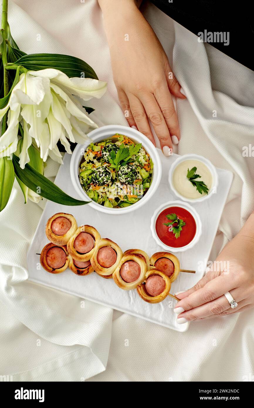Weibliche Hand nimmt appetitliche goldbraune Würstchen auf Spießen, serviert auf Teller mit Gemüsesalat und Saucen gegen weiße Tischdecke im Hintergrund Stockfoto