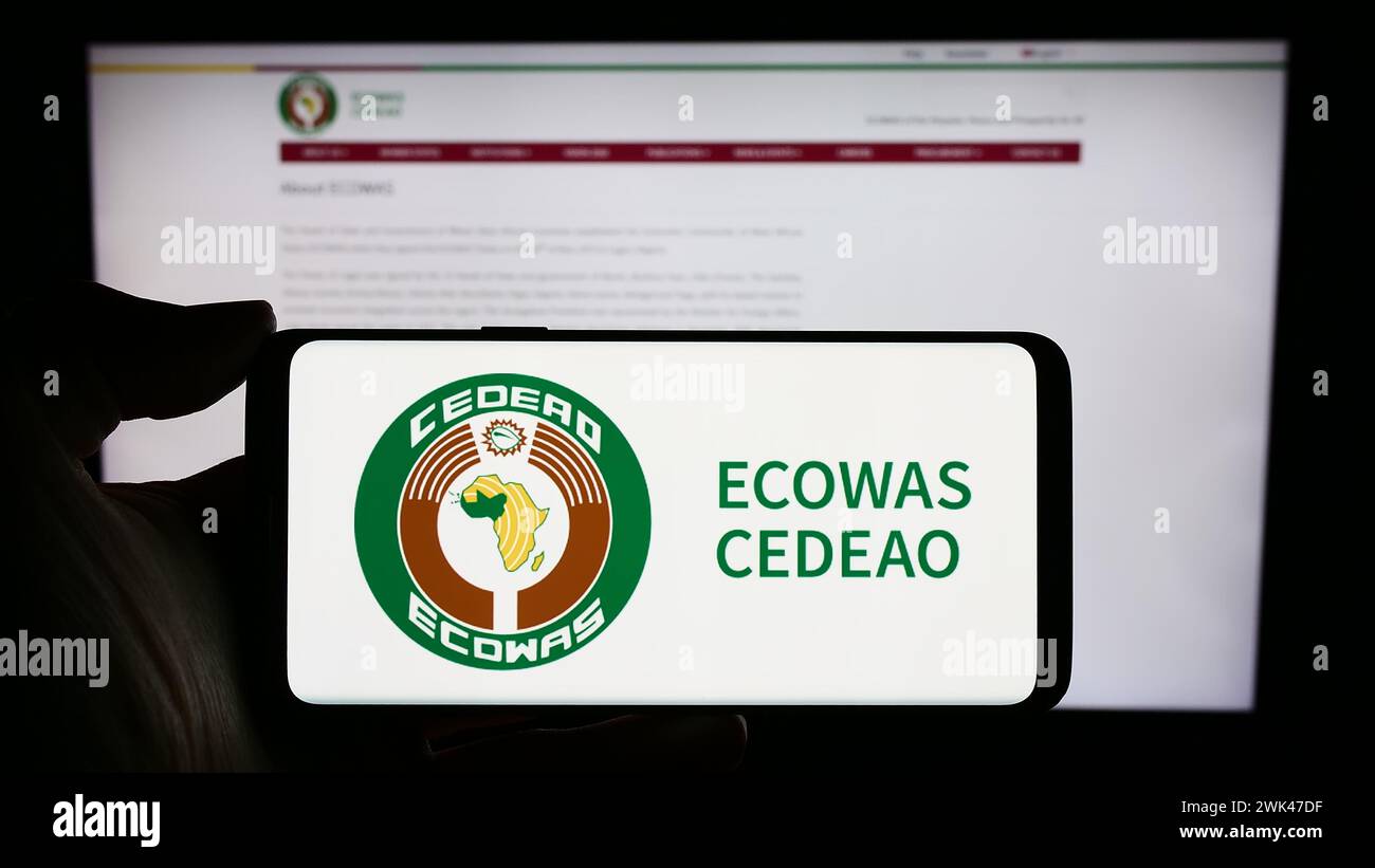 Person, die ein Mobiltelefon mit dem Logo der Wirtschaftsgemeinschaft westafrikanischer Staaten (ECOWAS) vor der Webseite hält. Schwerpunkt auf dem Display des Telefons. Stockfoto