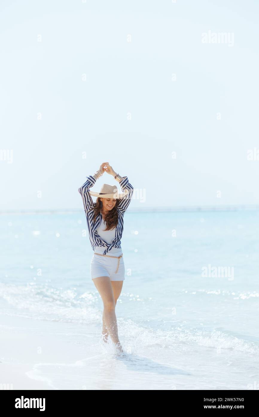 Porträt einer glücklichen modernen 40-jährigen Frau an der Küste des Ozeans mit Strohhut, die läuft. Stockfoto