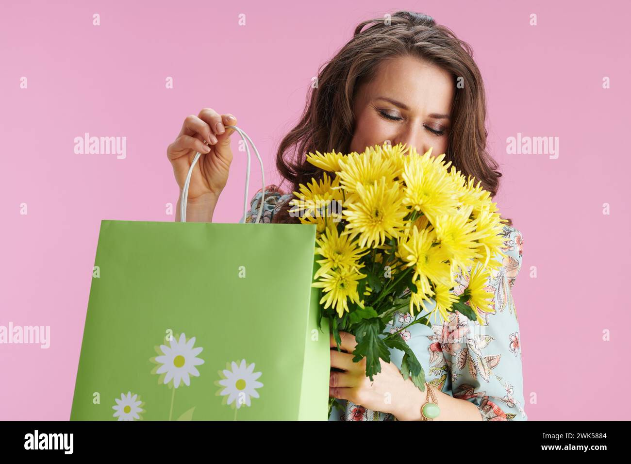 Glückliche trendige 40 Jahre alte Frau mit langen welligen brünetten Haaren mit gelben Chrysanthemen Blumen und grüner Einkaufstasche auf rosa Hintergrund. Stockfoto