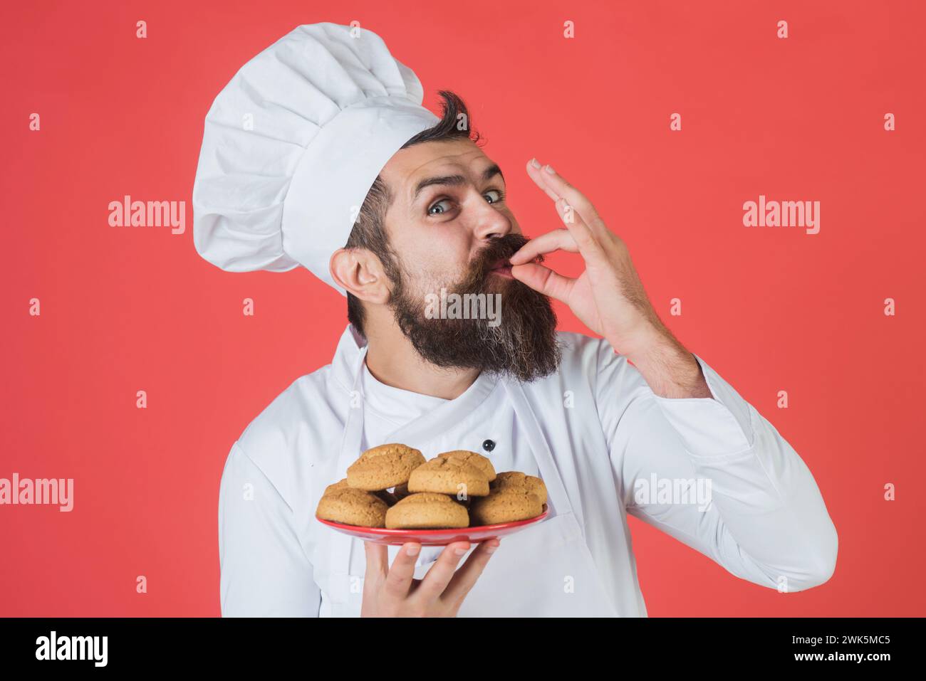 Bärtiger Mann in Küchenuniform mit einem Teller Haferflockenkekse, auf dem Zeichen lecker zu sehen ist. Gutaussehender Koch, Koch oder Bäcker mit Geste zur Geschmackszustimmung. Männlicher Koch Stockfoto