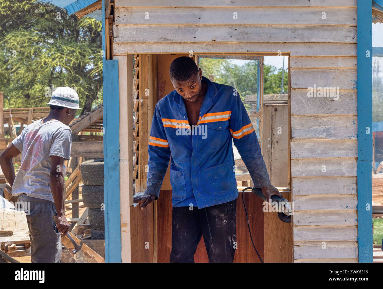 Zwei afroamerikanische Arbeiter bauen ein wendy-Haus, Holzschuppen im Freien, einer benutzt einen Winkelschleifer, um das Fenster zu glätten, der andere hämmert A Stockfoto