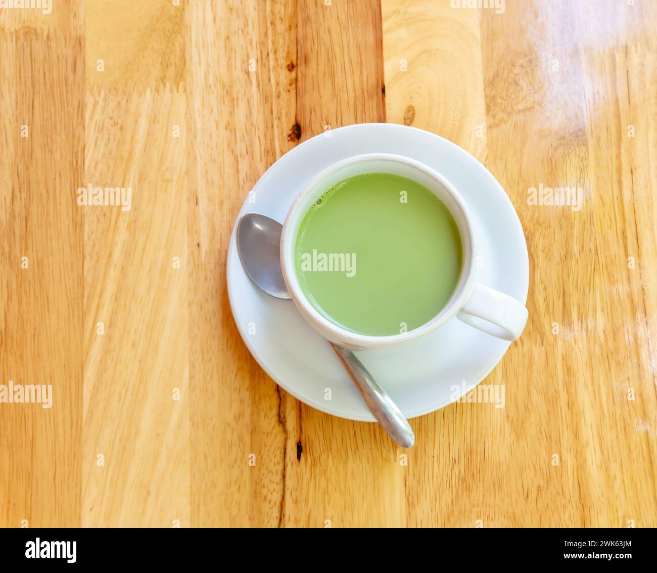 Blick von oben auf heiße Kondensmilch Matcha grüner Tee wird in weißer Tasse und kleiner Teller mit kleinem Teelöffel auf braunem Wooken Hintergrund serviert. Stockfoto