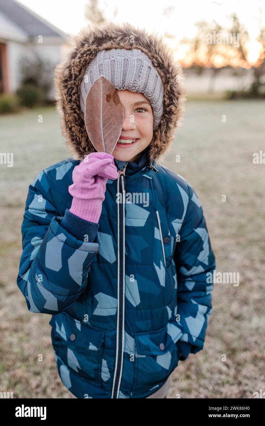 Kleines Mädchen, das einen geschwollenen Wintermantel trägt und ein gefrorenes Blatt hochhält Stockfoto