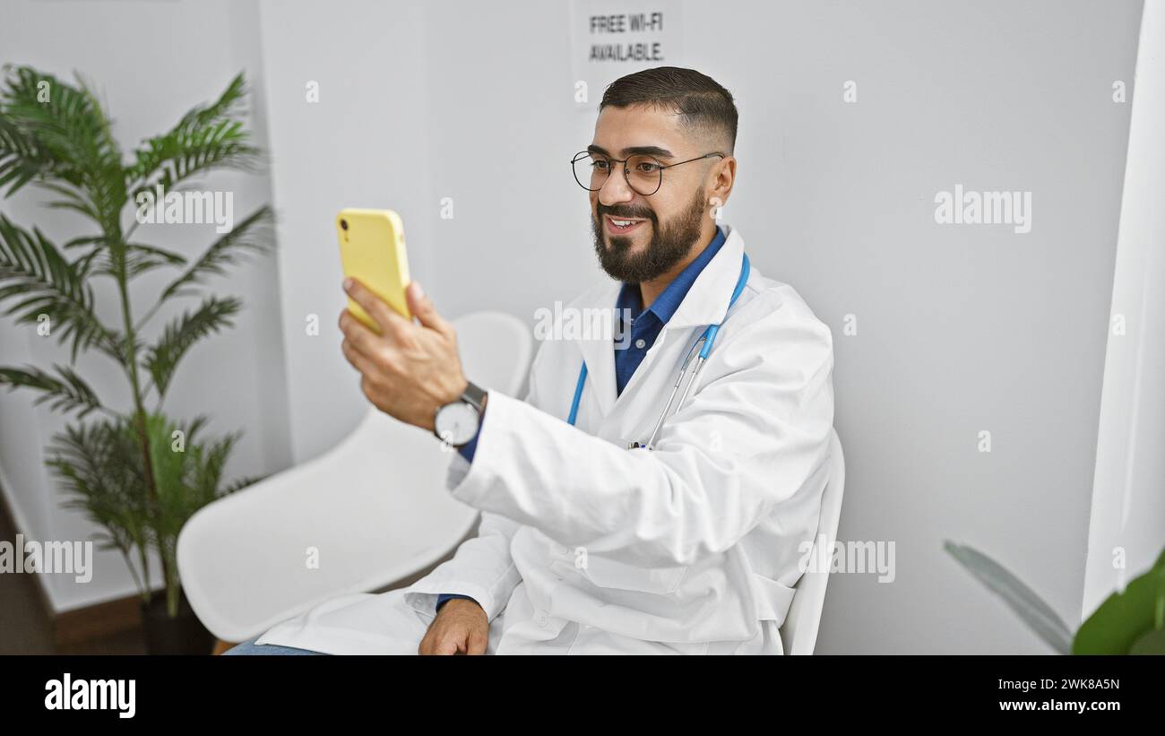 Ein lächelnder bärtiger Mann in einem weißen Mantel macht ein Selfie in einem modernen Raum mit einer Pflanze und einem wlan-Schild Stockfoto