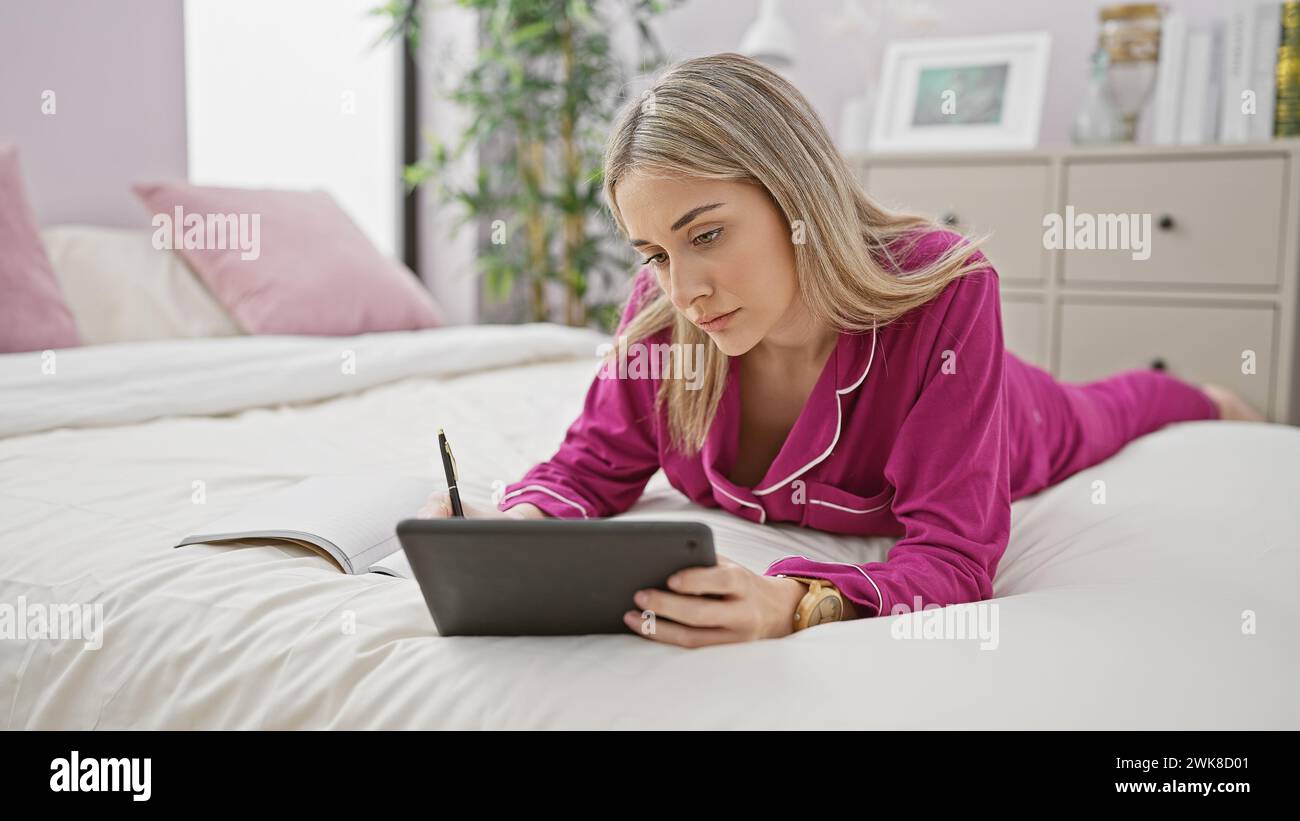 Eine junge Frau im Schlafanzug, die ein Tablet benutzt und in ein Notizbuch schreibt, während sie auf einem Bett in einer gemütlichen Schlafzimmerumgebung liegt. Stockfoto