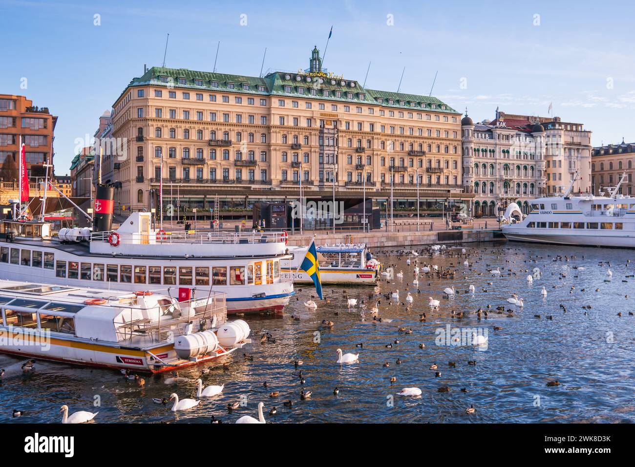 Sightseeing Boot in Stockholm, in der Nähe des Grand Hotels. Helles Sonnenlicht, Winter, alte Gebäude. Schwedische Flagge Stockfoto