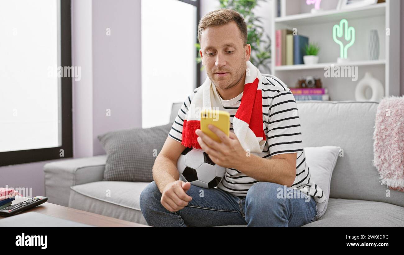 Leidenschaftlicher Fußballfan, junger kaukasischer Mann, der in intensive Wettkämpfe vertieft ist und per Smartphone vom gemütlichen Wohnzimmer-Sofa aus wettete Stockfoto