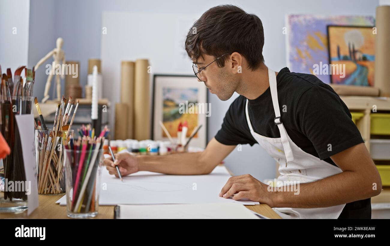 Der talentierte junge hispanische Künstler, der in einem lebendigen Kunststudio voll und ganz in das Zeichnen mit Bleistift eingetaucht ist und rohe Talente zwischen Pinseln und Leinwand zeigt Stockfoto