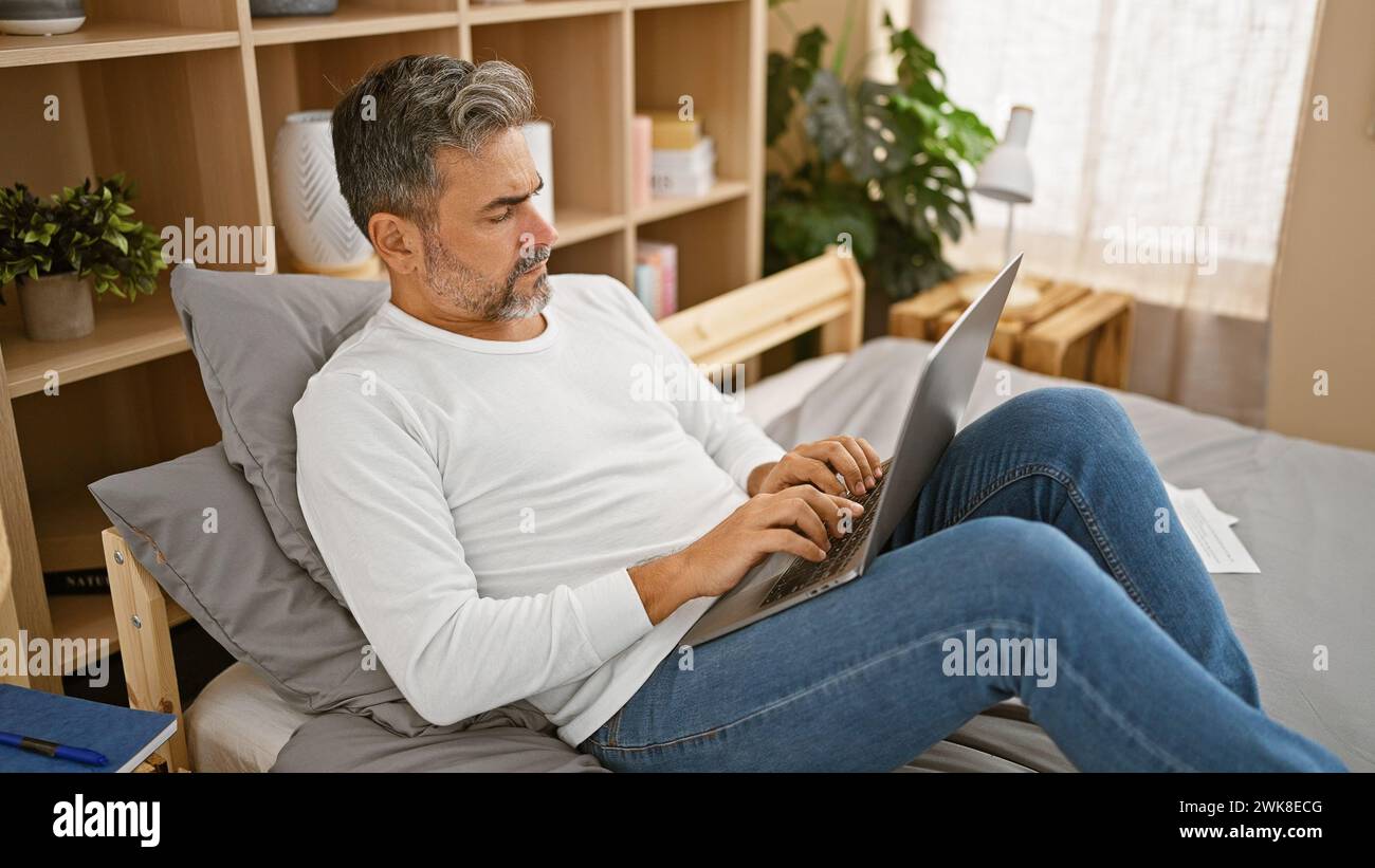 Entspannte Morgenstimmung, attraktiver junger hispanischer Mann, grauhaarig und bärtig, bequem auf einem gemütlichen Bett sitzend und entspannend im Internet surfen Stockfoto Entspannte Morgenstimmung, attraktiver junger hispanischer Mann, grauhaarig und bärtig, bequem auf einem gemütlichen Bett sitzend und entspannend im Internet surfen Stockfoto