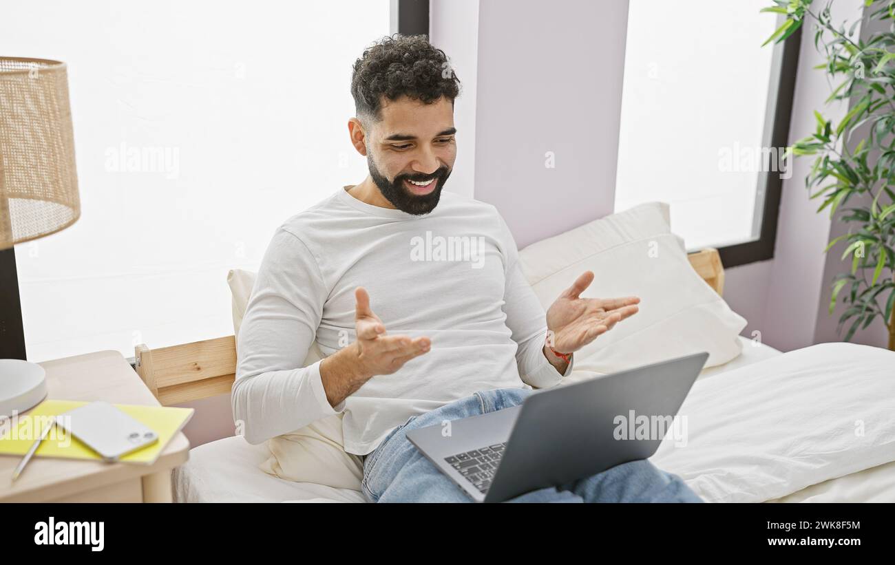 Lächelnder bärtiger Mann, der einen Laptop benutzt und in einem hellen Schlafzimmer gestikuliert. Stockfoto